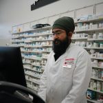 Asian male pharmacist in a lab coat working at a pharmacy counter with shelves of medicine.